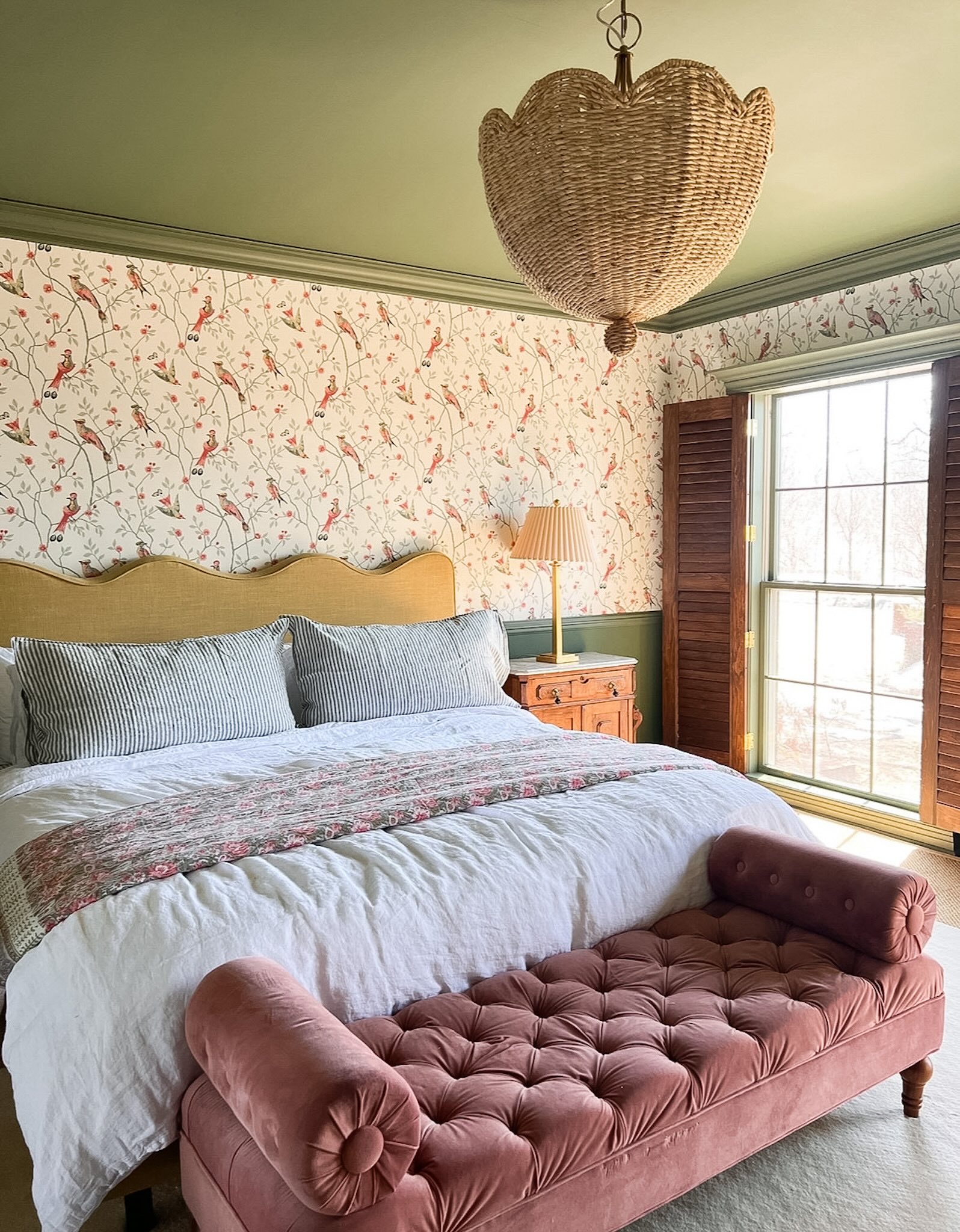 primary bedroom with scalloped headboard, rattan pendant light, pink tufted velvet bench, and bird wallpaper