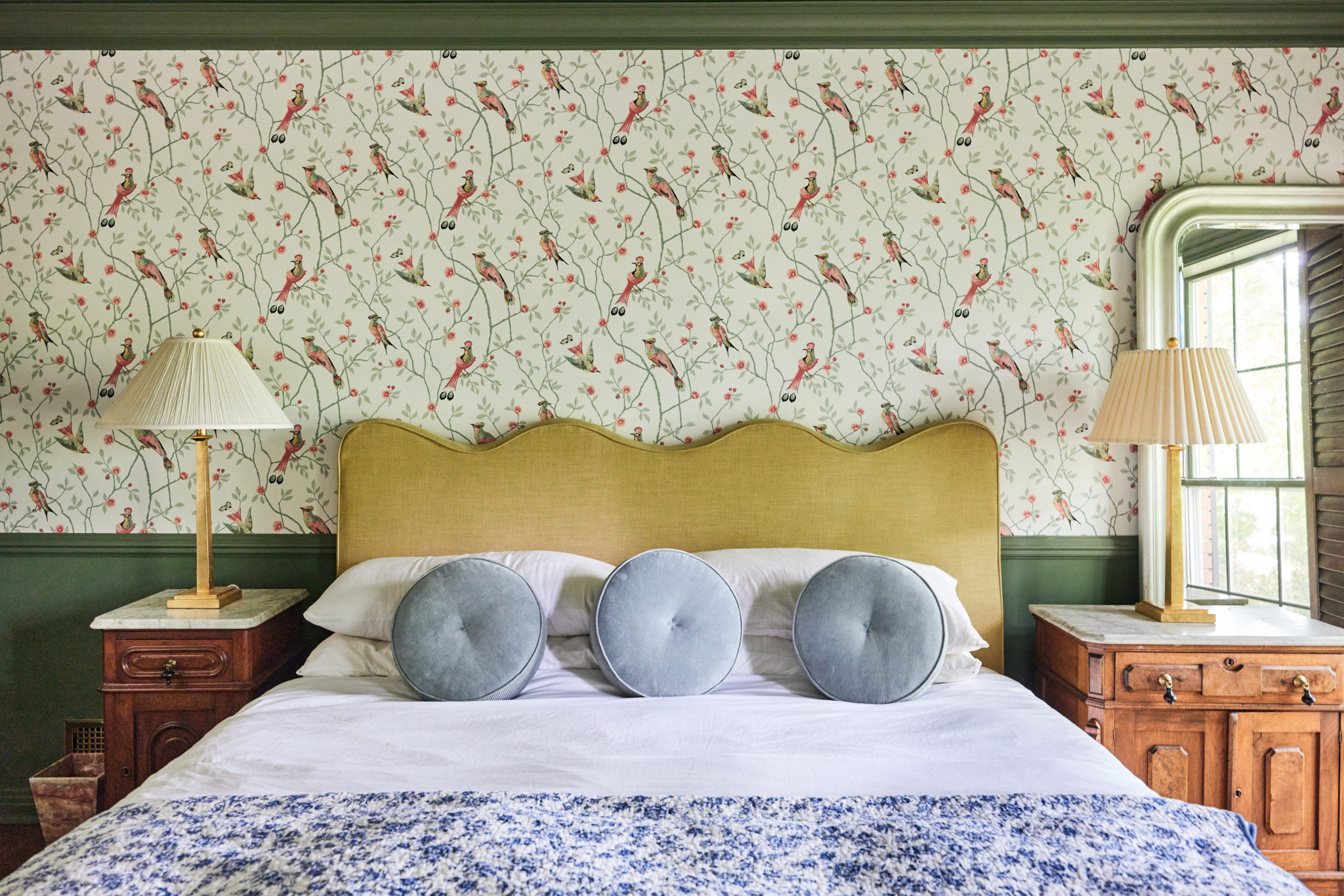 A main bedroom with a scalloped headboard, antique wood and marble nighstands, green trim, and bird wallpaper