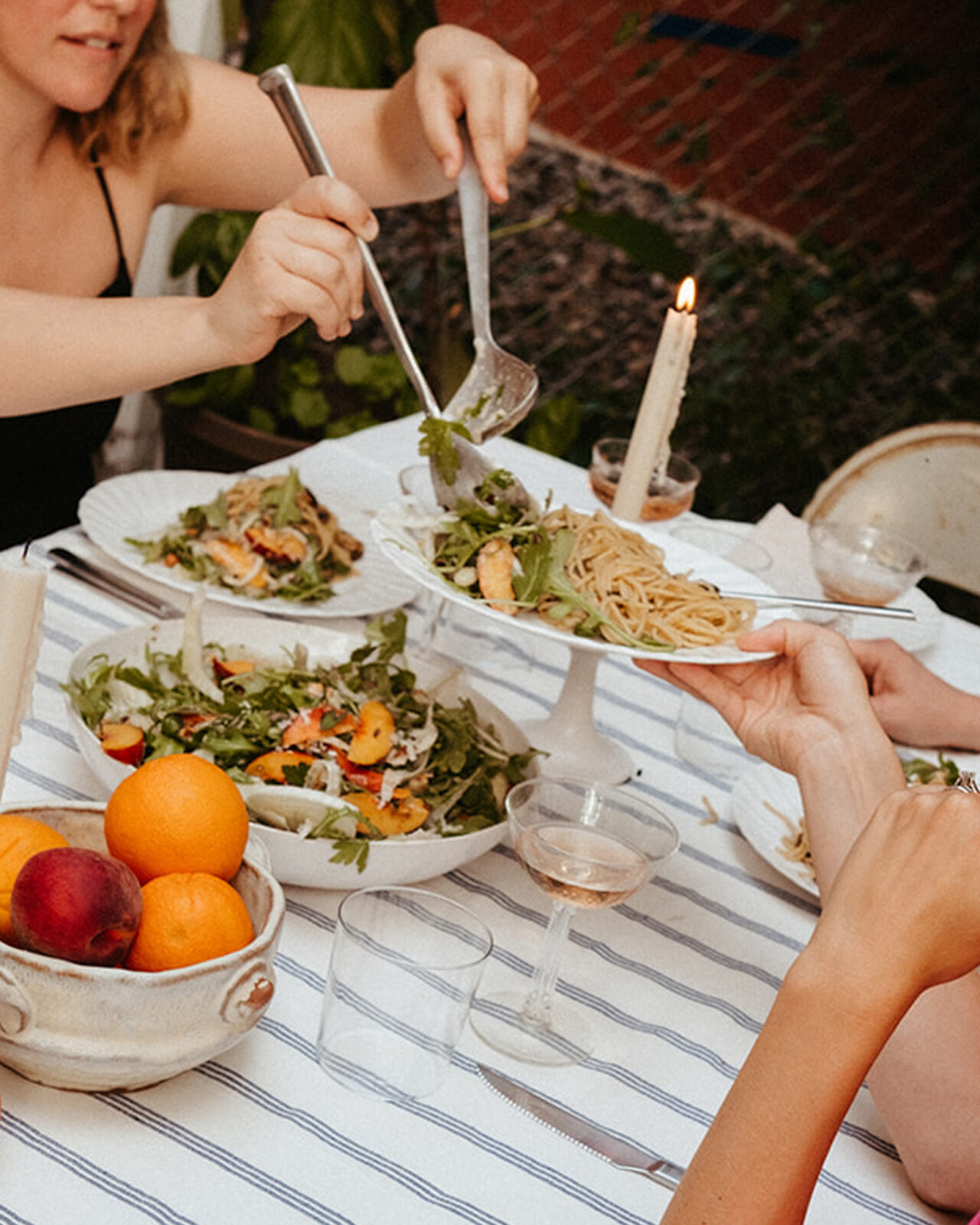 A table is set for a summer dinner party, with an arugula and peach salad and champagne coupes on top of a striped cotton tablecloth