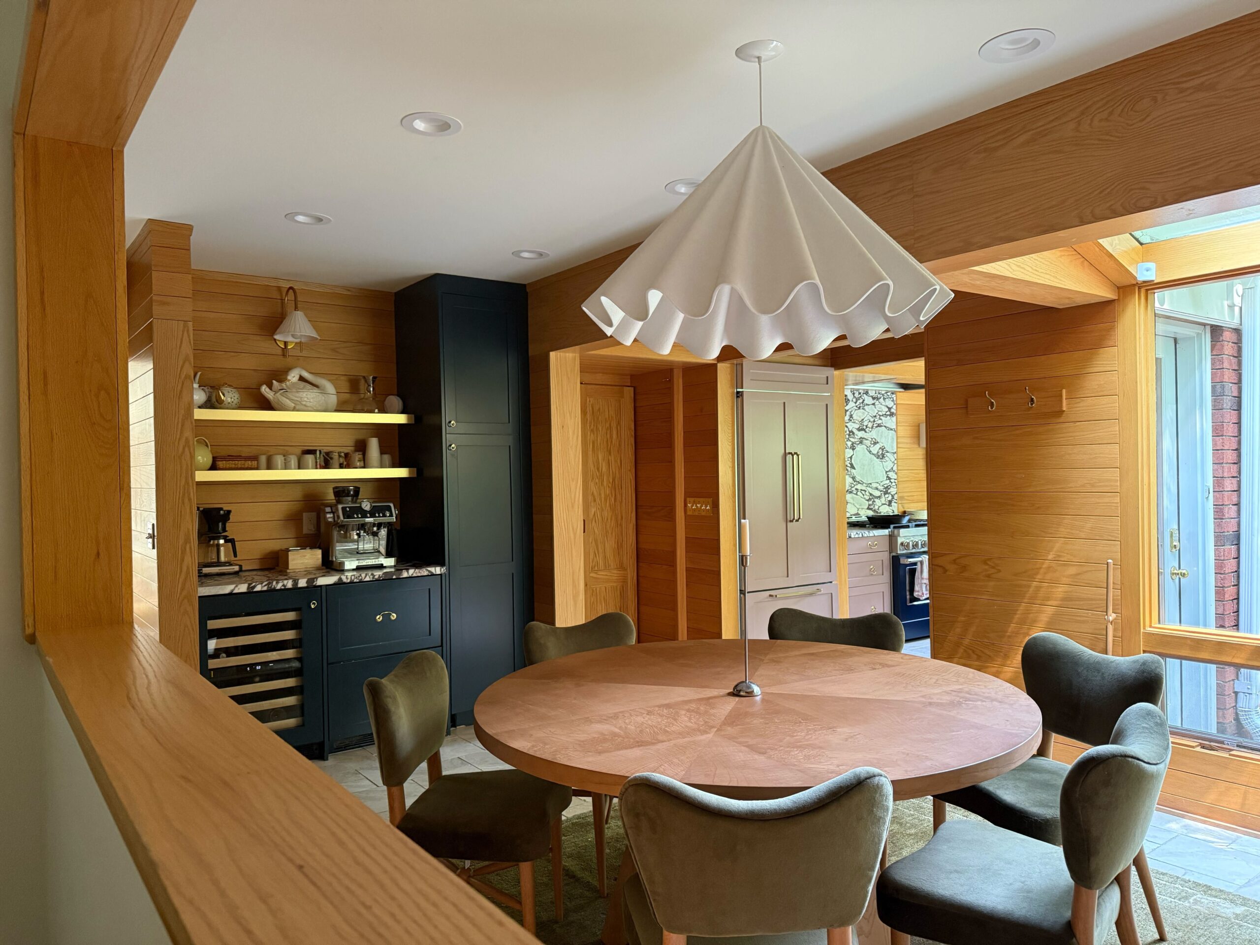 In an oak-paneled eat-in kitchen sits a round burl wood dining table surrounded by green upholstered dining chairs from Pierce & Ward by West Elm