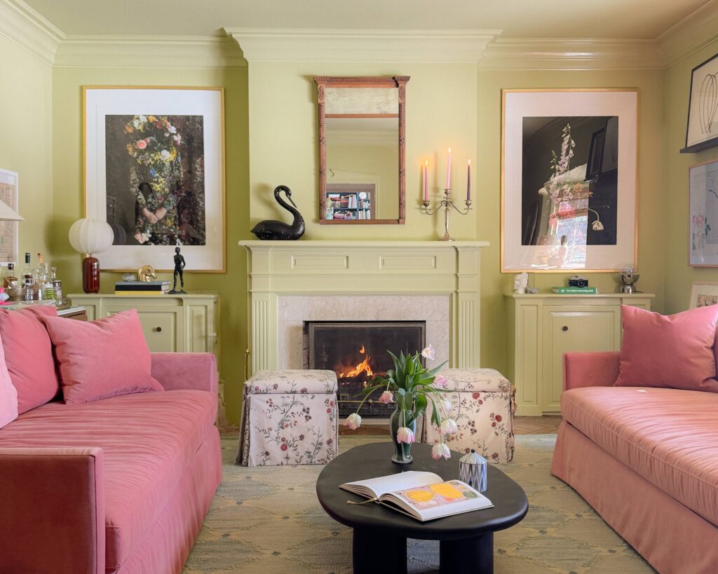 A green family room with pink velvet sofas, skirted ottomans, and a black oval coffee table.