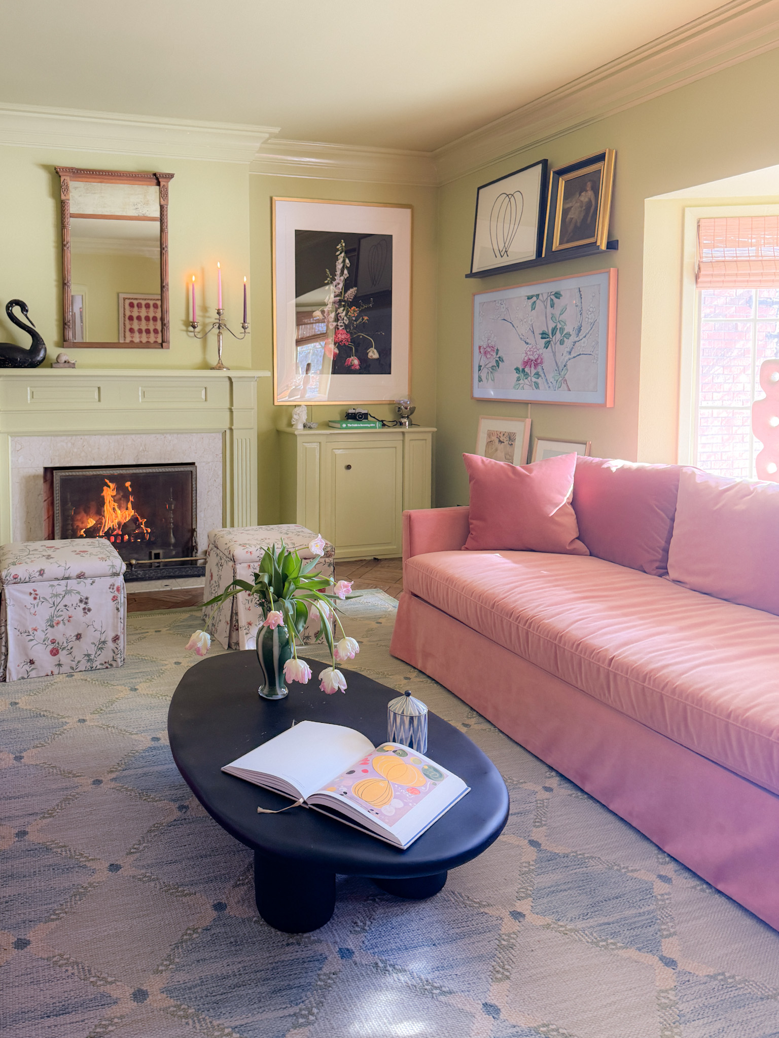 A green family room with pink velvet sofas, skirted ottomans, and a black oval coffee table.