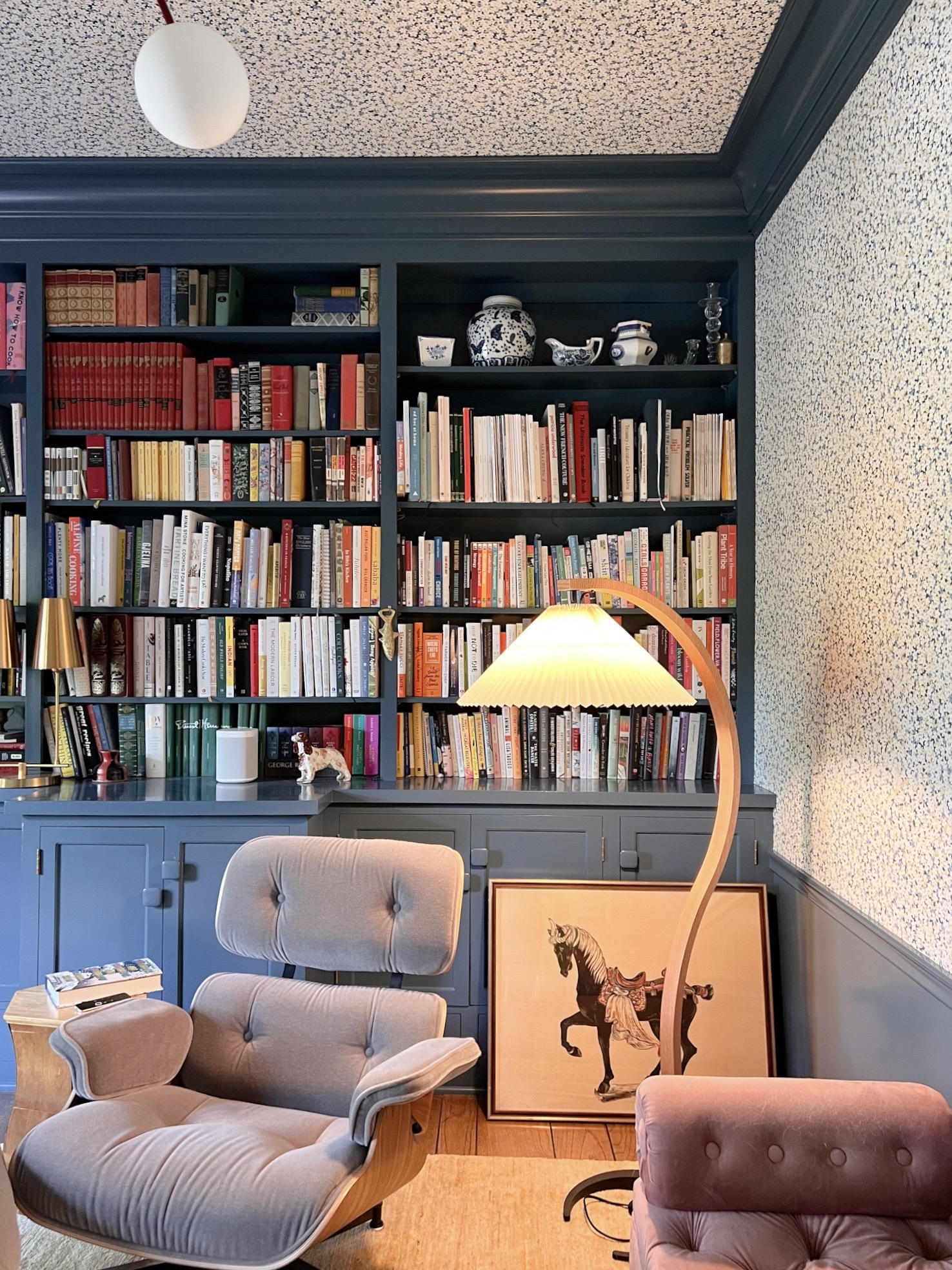 an office with blue built-in bookshelves, floral wallpaper, and a velvet eames chair